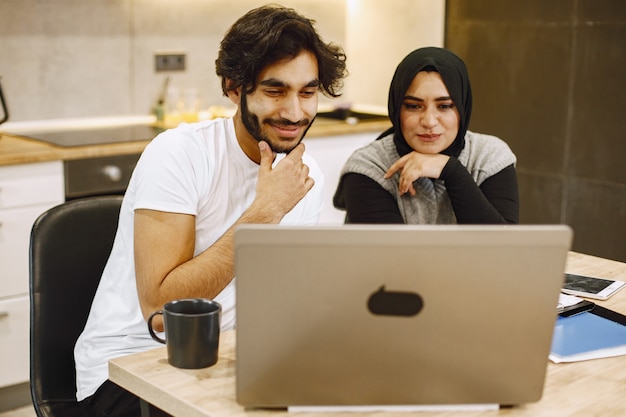 Schönes junges Paar, das einen Laptop verwendet, in ein Notizbuch schreibt und zu Hause in einer Küche sitzt. Arabisches Mädchen mit schwarzem Hidjab.