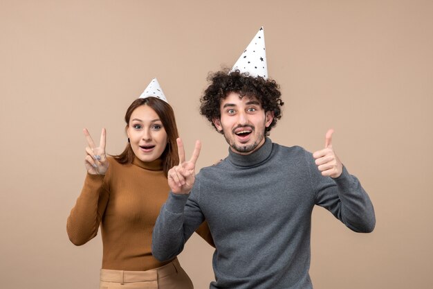 Schönes junges Paar tragen Neujahrshut lächelndes Mädchen zeigt Siegesgeste lustiger Kerl, der ok auf grau zeigt