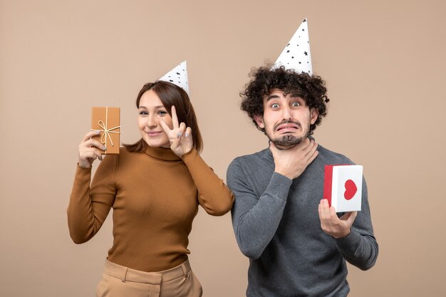 Schönes junges Paar tragen Neujahrshutmädchen mit Geschenk, das Siegesgeste angespannten Kerl mit Herz auf Grau macht