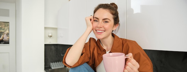 Schönes junges weibliches Modell hält eine rosa Tasse, sitzt in der Küche und lächelt bei der Kamerafrau