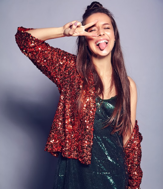 Schönes kaukasisches lächelndes Hipster-Brünettenfrauenmodell in der hellen glänzenden reflektierenden sommerlichen stilvollen Jacke und im grünen Kleid, die auf Grau aufwerfen. Friedenszeichen zeigen