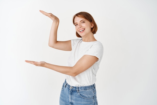 Schönes kaukasisches Mädchen mit natürlichem Schönheitsgesicht, das ein großes Objekt auf dem Kopierraum zeigt, eine große Schachtel mit den Händen formen und auf einer weißen Wand stehen