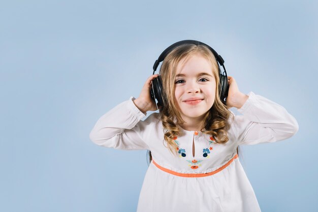 Schönes kleines Mädchen, welches die Musik auf Kopfhörer gegen blauen Hintergrund genießt