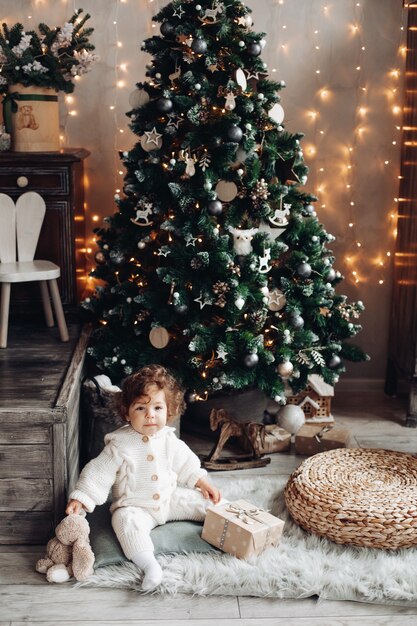 Schönes Kleinkind mit einem Plüschhaustier und einem Geschenk, das auf dem Teppich nahe Weihnachtsbaum sitzt