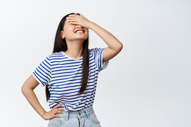 Schönes koreanisches Mädchen, das ihre Augen überrascht, lächelt aus Vorfreude, wartet auf etwas mit verbundenen Augen und steht auf Weiß.