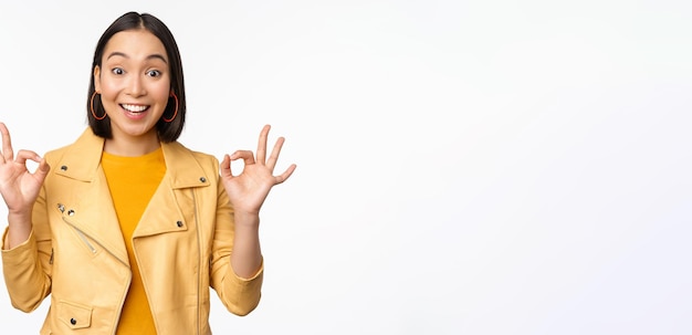 Schönes koreanisches mädchen mit glücklichem gesicht, das zufrieden aussieht und zeigt, dass es okay ist, geste zu empfehlen, shop zu mögen und etw zu genehmigen, guter weißer hintergrund von ausgezeichneter qualität