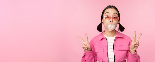 Kostenloses Foto schönes koreanisches mädchen mit sonnenbrille, das kaugummiblasen bläst und friedenszeichen zeigt, die über rosa hintergrund stehen