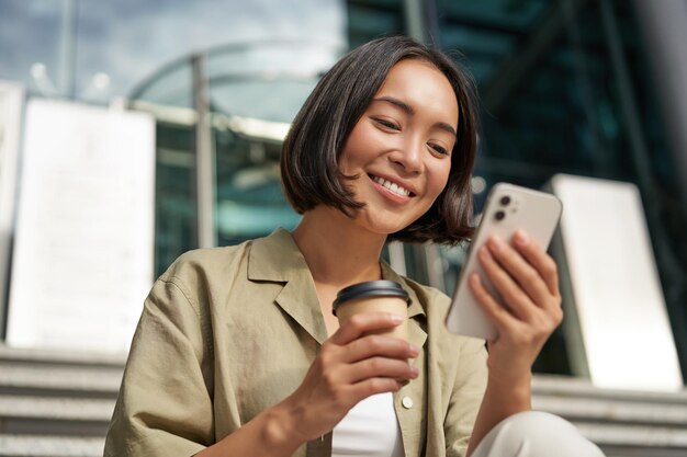 Schönes lächelndes asiatisches Mädchen, das mit dem Handy Kaffee trinkt und auf der Treppe vor der jungen Frau sitzt