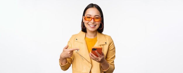 Schönes lächelndes asiatisches Mädchen mit Sonnenbrille, das mit dem Finger auf das Smartphone zeigt, das den App Store auf dem Handy zeigt, das über weißem Hintergrund steht