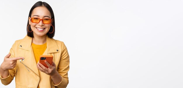 Schönes lächelndes asiatisches Mädchen mit Sonnenbrille, das mit dem Finger auf das Smartphone zeigt, das den App Store auf dem Handy zeigt, das über weißem Hintergrund steht