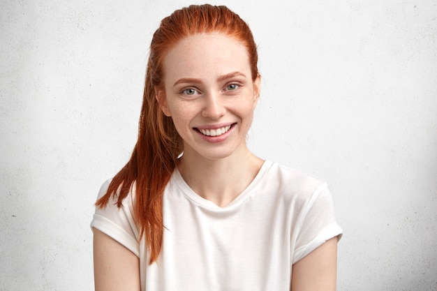 Schönes lächelndes entzücktes junges weibliches Modell mit Ingwerhaar und Sommersprossen im Gesicht, gekleidet in lässigem weißem T-Shirt, drückt positive Emotionen aus