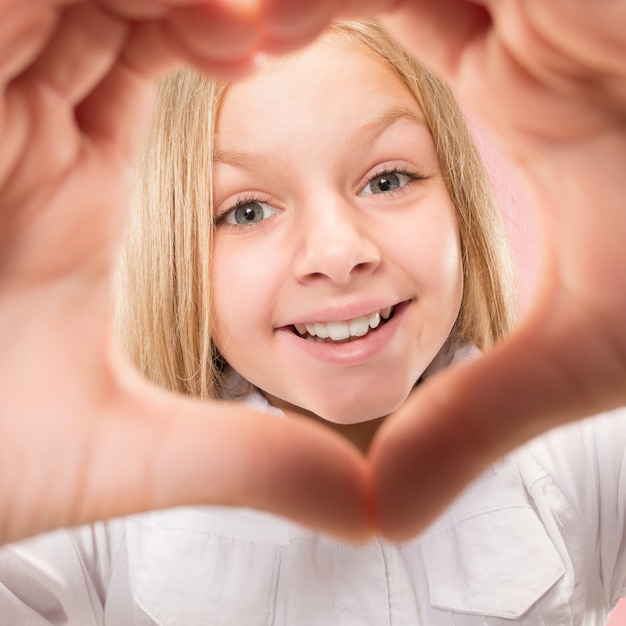 Kostenloses Foto schönes lächelndes jugendlich mädchen macht die form eines herzens mit ihren händen auf dem rosa hintergrund. liebesgeste des hübschen kleinen kindes.