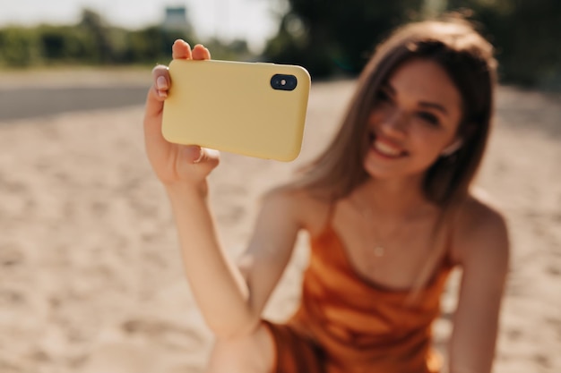 Schönes lächelndes Mädchen mit dunkler Frisur macht Selfie im Freien bei Sonnenschein, während sie sich am Strand ausruht Telefon im Fokus im Vordergrund Mädchen im Hintergrund unscharf