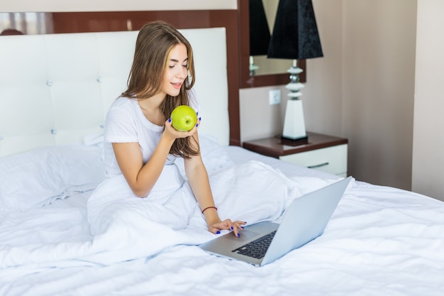 Schönes lächelndes Mädchen sitzt früh morgens im Bett, isst einen Apfel und lächelt mit einem Laptop