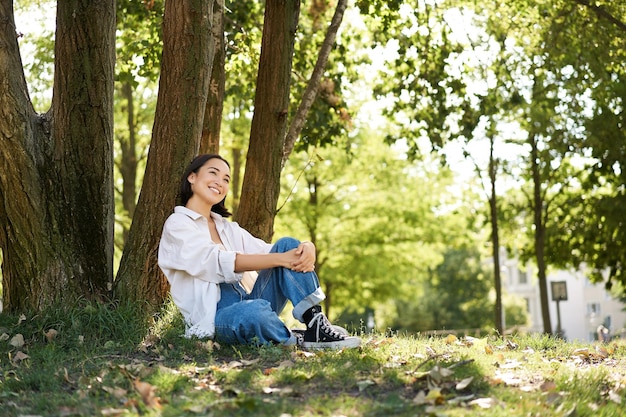 Schönes lächelndes Mädchen sitzt in der Nähe von Bäumen im Park und genießt die Natur im Freien, entspannt und ruht sich auf frischem aus