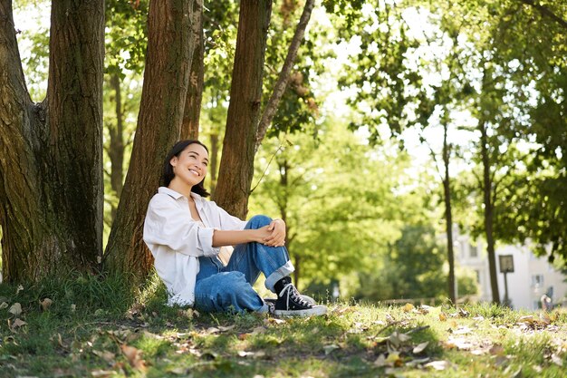 Schönes lächelndes Mädchen sitzt in der Nähe von Bäumen im Park und genießt die Natur im Freien, entspannt und ruht sich auf frischem aus