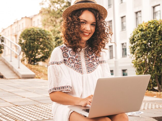 Schönes lächelndes Modell mit Afro kräuselt Frisur im weißen Kleid und im Hut des Sommerhippies.