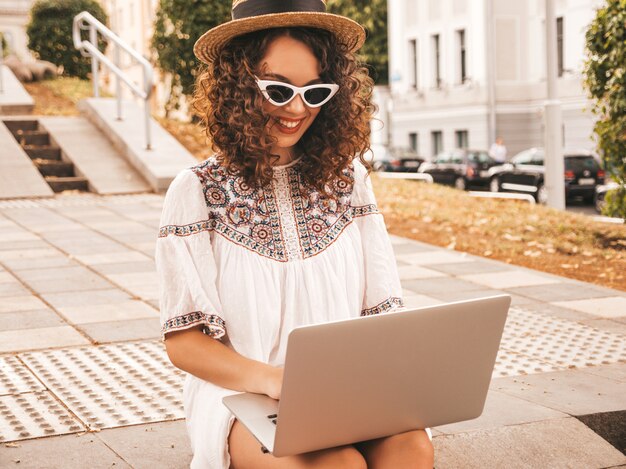 Schönes lächelndes Modell mit Afro kräuselt Frisur im weißen Kleid und im Hut des Sommerhippies.