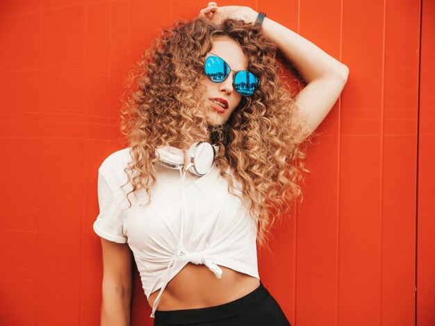 Schönes lächelndes Modell mit Afro-Lockenfrisur gekleidet in Sommer-Hipster-Kleidung. Sexy sorgloses Mädchen, das nahe rote Wand im Freien aufwirft. Lustige und positive Frau, die Spaß in der Sonnenbrille hat