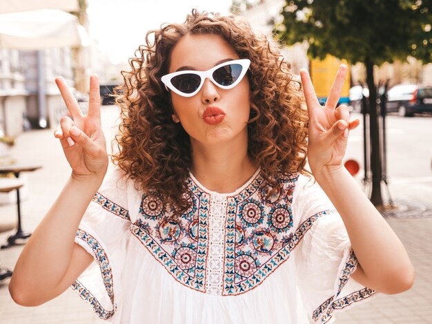 Schönes lächelndes Modell mit Afrolockenfrisur kleidete im weißen Kleid des Sommerhippies an.