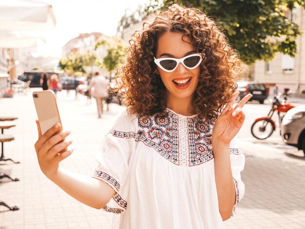 Schönes lächelndes Modell mit Afrolockenfrisur kleidete im weißen Kleid des Sommerhippies an.