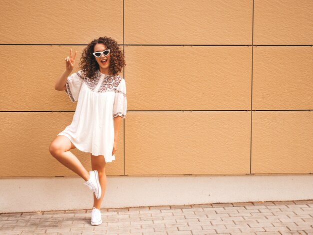 Schönes lächelndes Modell mit Afrolockenfrisur kleidete im weißen Kleid des Sommerhippies an.