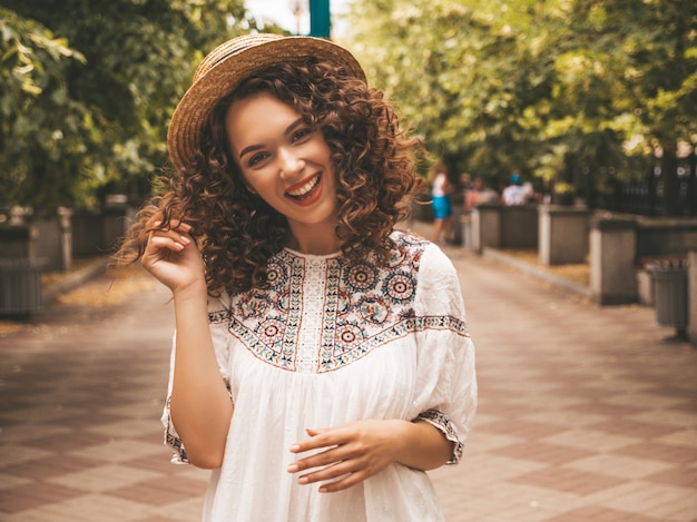 Schönes lächelndes Modell mit Afrolockenfrisur kleidete im weißen Kleid des Sommerhippies an.