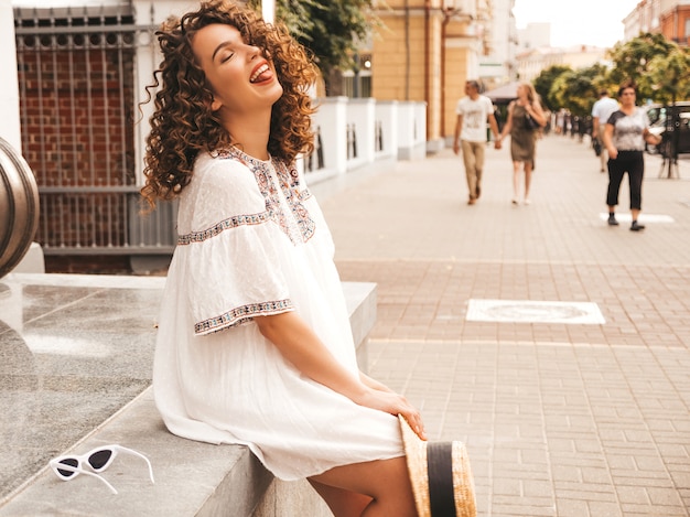 Schönes lächelndes Modell mit Afrolockenfrisur kleidete im weißen Kleid des Sommerhippies an.