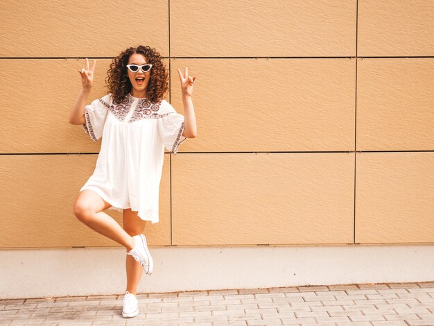 Schönes lächelndes Modell mit Afrolockenfrisur kleidete im weißen Kleid und in der Sonnenbrille des Sommerhippies an