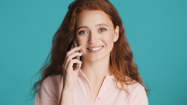 Schönes lächelndes rothaariges Mädchen, das am Telefon spricht und glücklich in die Kamera blickt, vor buntem Hintergrund