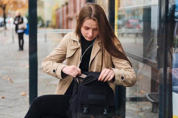 Schönes lässiges Mädchen in Kopfhörern, das etwas im Rucksack sieht und auf öffentliche Verkehrsmittel an der Bushaltestelle im Freien wartet