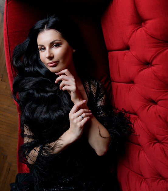 Schönes langhaariges brünettes kaukasisches Mädchen schaut gerade und liegt auf dem roten Sofa