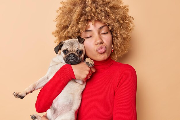 Schönes, lockiges weibliches Modell umarmt Pug-Hund mit Liebe, hat die Augen geschlossen, schmollt die Lippen und drückt die Fürsorge für ihr Lieblingshaustier aus, das über braunem Hintergrund isoliert ist Menschen, Haustiere und Freundschaftskonzept