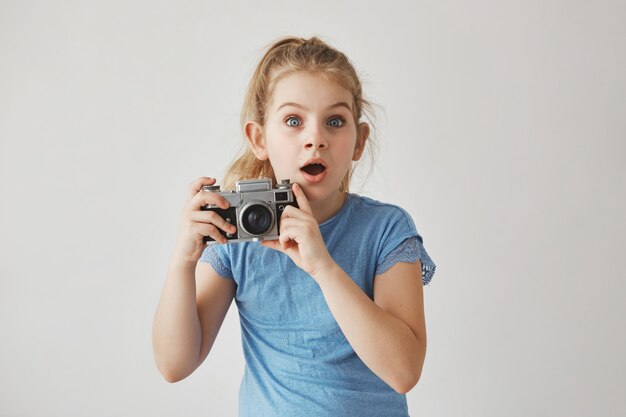 Schönes lustiges kleines Mädchen mit blondem Haar und blauen Augen, die Fotokamera in Händen halten und gerade mit erschrockenem Ausdruck schauen.