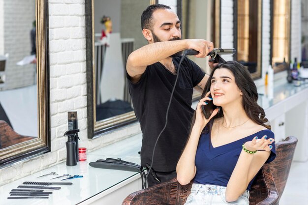 Schönes Mädchen, das am Telefon spricht, während der Friseur ihr Haar formt