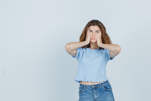 Schönes mädchen, das hände auf wangen in t-shirt, jeans drückt und müde, vorderansicht schaut.