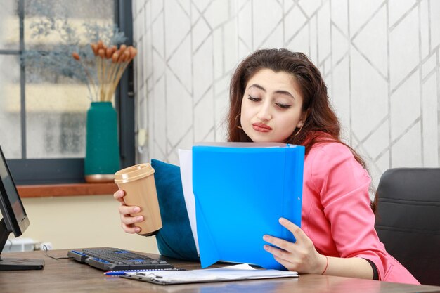 Schönes Mädchen, das hinter dem Schreibtisch sitzt und Kaffee trinkt, während es ihre Notizen liest Hochwertiges Foto
