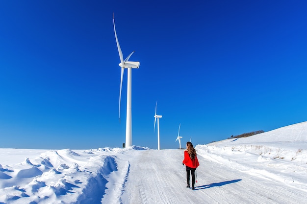 Schönes Mädchen, das in der Winterlandschaft des Himmels und der Winterstraße mit Schnee und rotem Kleid und Windkraftanlage geht