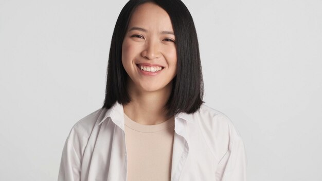 Schönes Mädchen, das Kamera betrachtet und aufrichtig lächelnde asiatische Frau, die über weißem Hintergrund steht