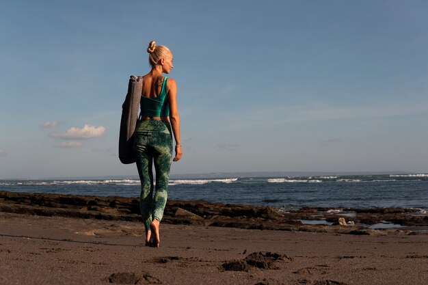 Schönes Mädchen, das mit Yogamatte am Strand spazieren geht