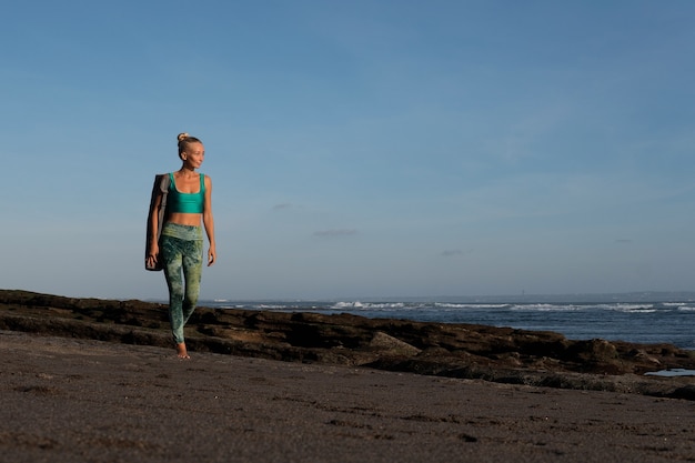 Schönes Mädchen, das mit Yogamatte am Strand spazieren geht