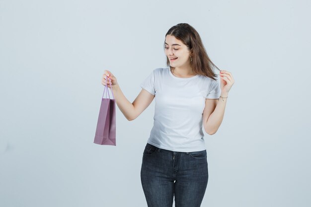 Schönes Mädchen, das Papiertüte im T-Shirt, in den Jeans und in der glücklichen Vorderansicht hält.