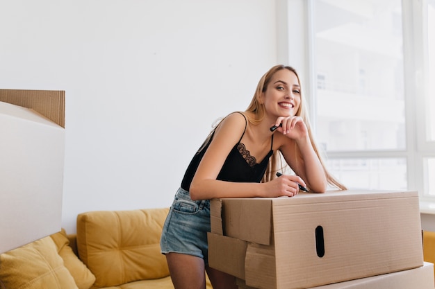 Schönes Mädchen, das Sachen packt, Karton etikettiert, Marker in der Hand hält, Umzug in neue Wohnung, Wohnung, Haus. Lächelnde Frau im Zimmer mit gelbem Sofa, sie trägt Jeansshorts, schwarzes Oberteil.