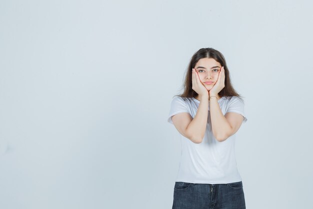 Schönes Mädchen, das Wangen mit den Händen im T-Shirt, in den Jeans hält und ernst schaut. Vorderansicht.