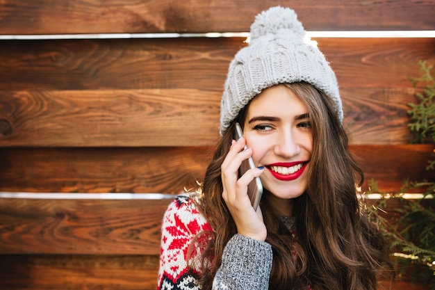 Schönes Mädchen des Nahaufnahmeporträts mit langen Haaren und schneeweißem Lächeln mit gestrickter Mütze auf Holz. Sie trägt einen warmen Pullover, telefoniert und lächelt zur Seite.