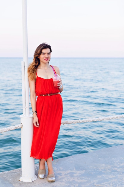 Schönes Mädchen im roten Kleid, das auf einem Pier steht und lächelt. Mädchen hat Ombre-Haare und hält eine Tasse mit kaltem Getränk und lila Strohhalm. Sie trägt hellbraune Espadrilles.