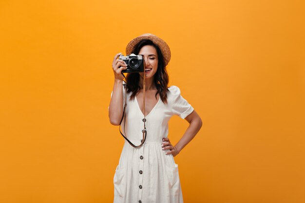 Schönes Mädchen im stilvollen Sommeroutfit macht Bild auf Retro-Kamera. Modische Frau im weißen langen Kleid und im lächelnden Hut.