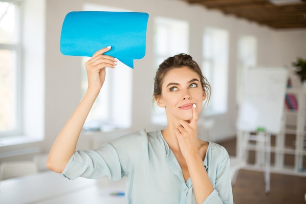 Schönes Mädchen in Bluse, das träumerisch beiseite schaut und blaues Papiernachrichtensymbol in der Hand in der Nähe des Kopfes hält, während es Zeit im Büro verbringt