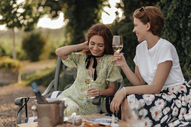 Schönes Mädchen in grüner Kleidung, das Glas mit Champagner hält und mit der Dame im Sommerblumenrock und hellem T-Shirt im Freien sitzt