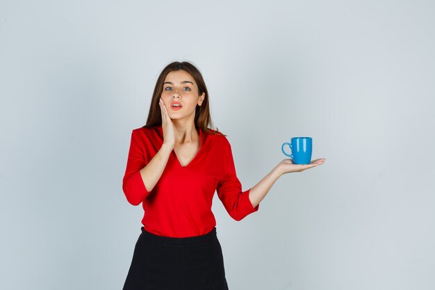 Schönes Mädchen in roter Bluse, schwarzer Rock, der Tasse hält und schockiert aussieht, Vorderansicht.